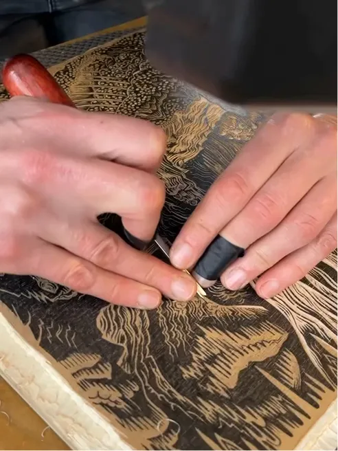 Hands cutting into an inked woodblock with a carving tool.